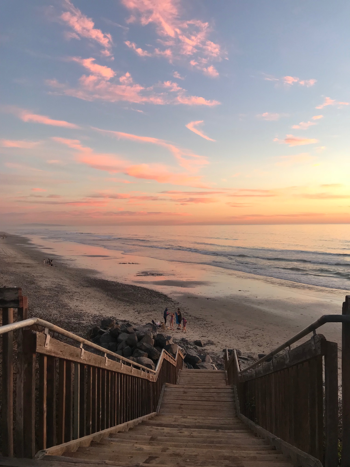 A wooden staircase descending to a quiet beach at sunset, with soft pink and gold clouds reflected on the wet sand.