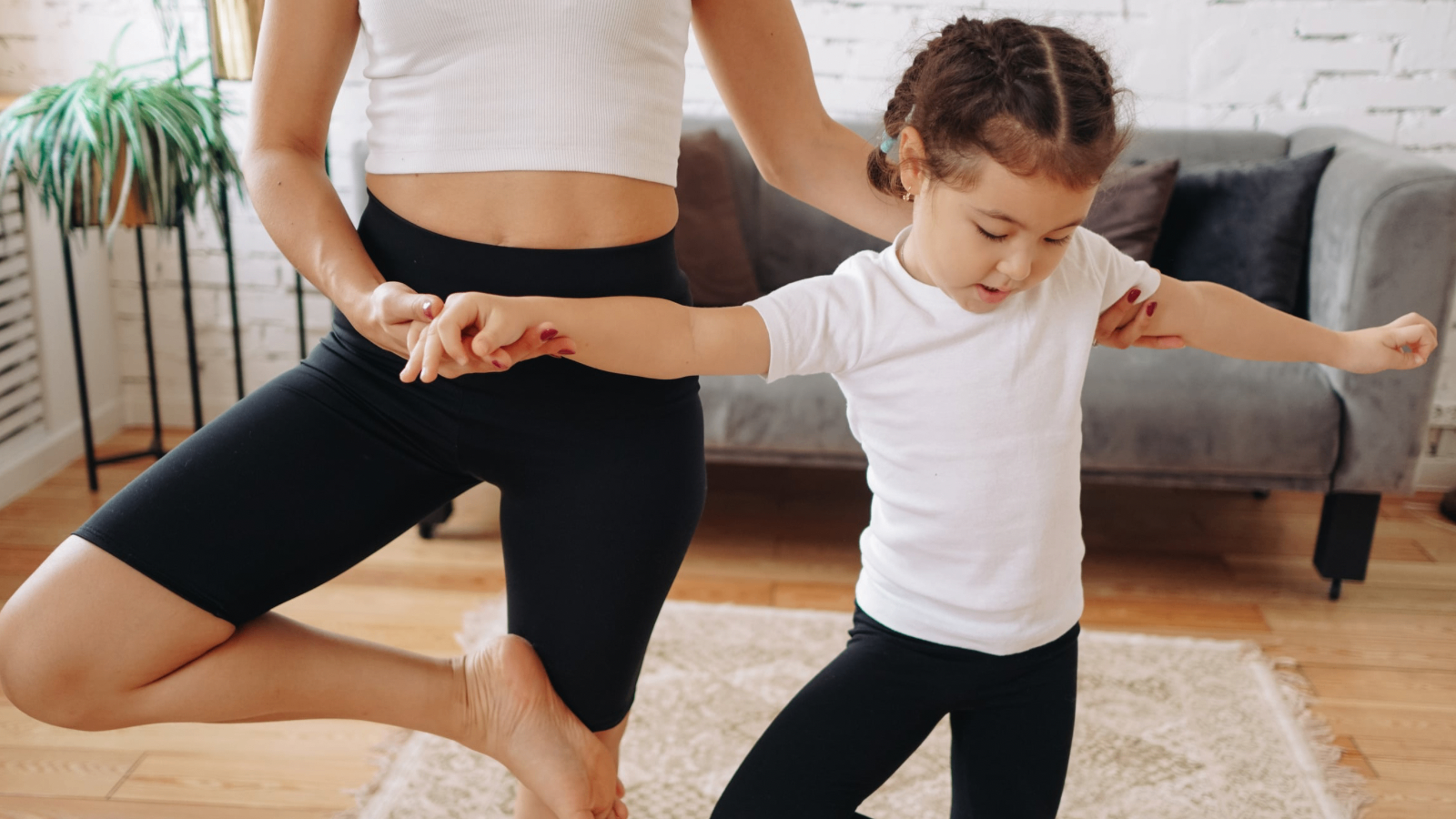 A mother and young daughter balancing together in a sunlit living room.