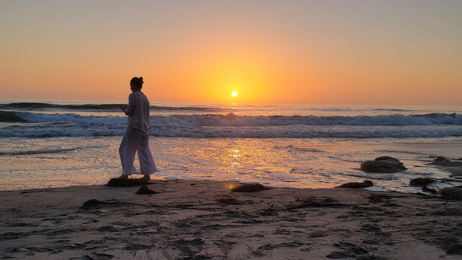Silhouette of a woman standing at the water's edge in stillness as the sun sets over the ocean, a quiet moment of embodied somatic awareness